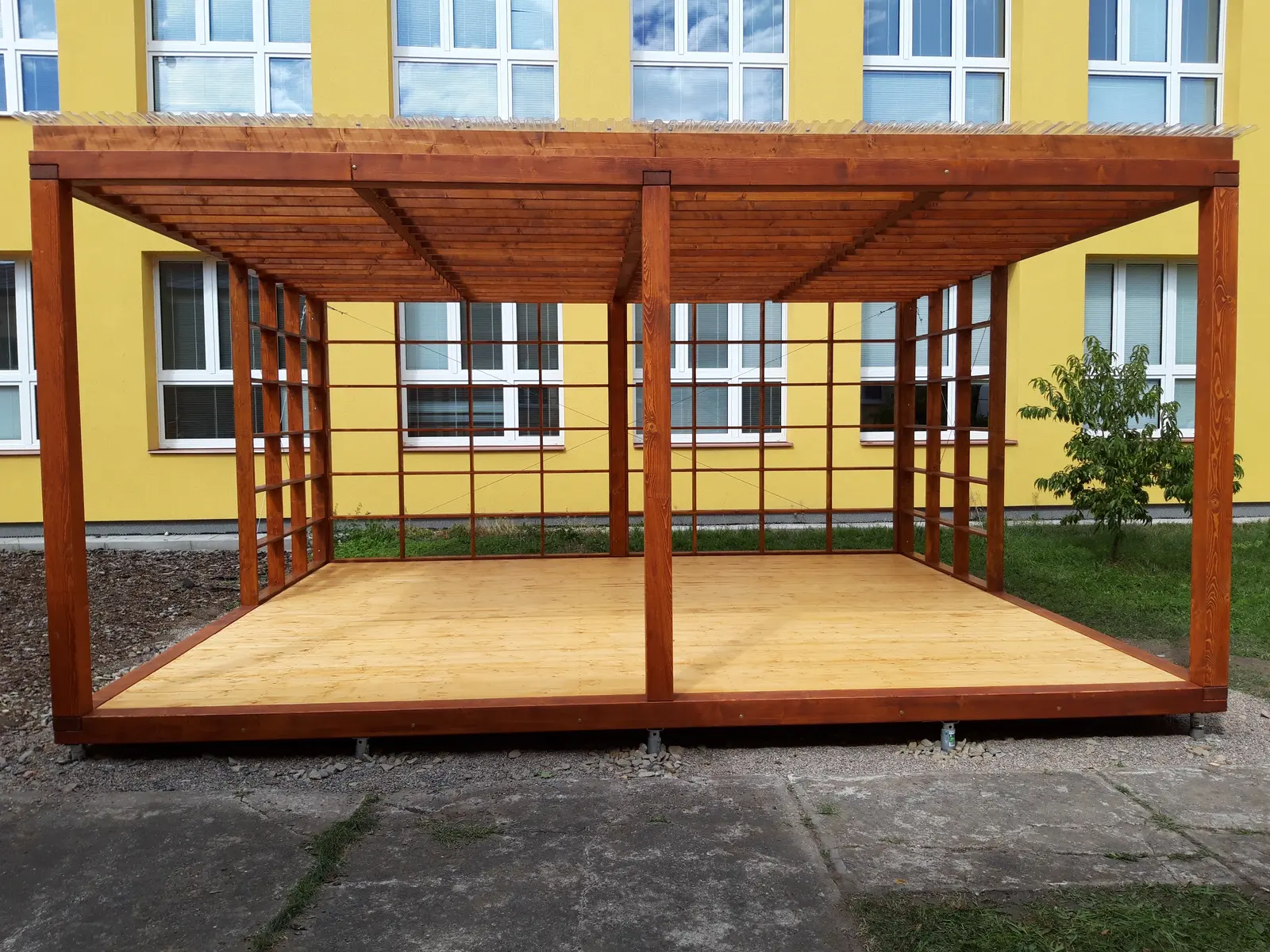 Dřevěná pergola s podlahou a mřížovými bočnicemi před školní budovou